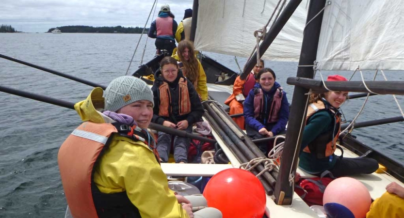 A group of students wearing life jackets sit inside a sailboat. Many of them are holding oars. 